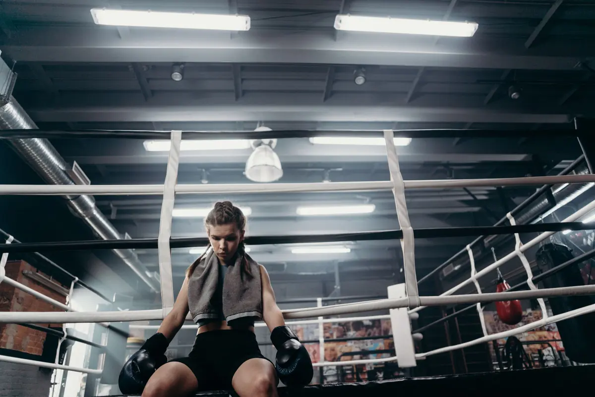 A woman in boxing gloves sits on the ring apron with a towel around her neck between rounds.