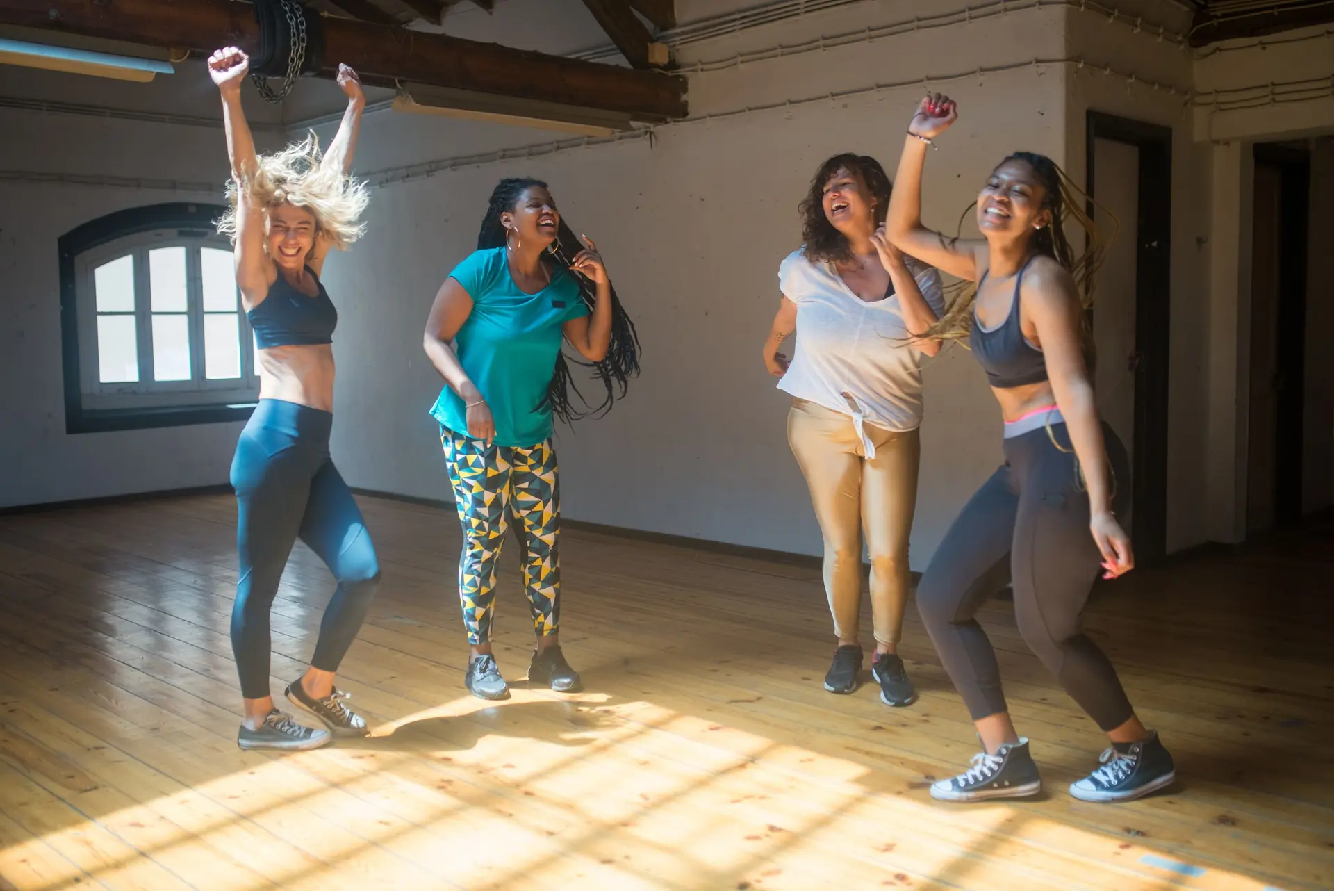 Four women laugh and dance together in the sunlight of a studio class.