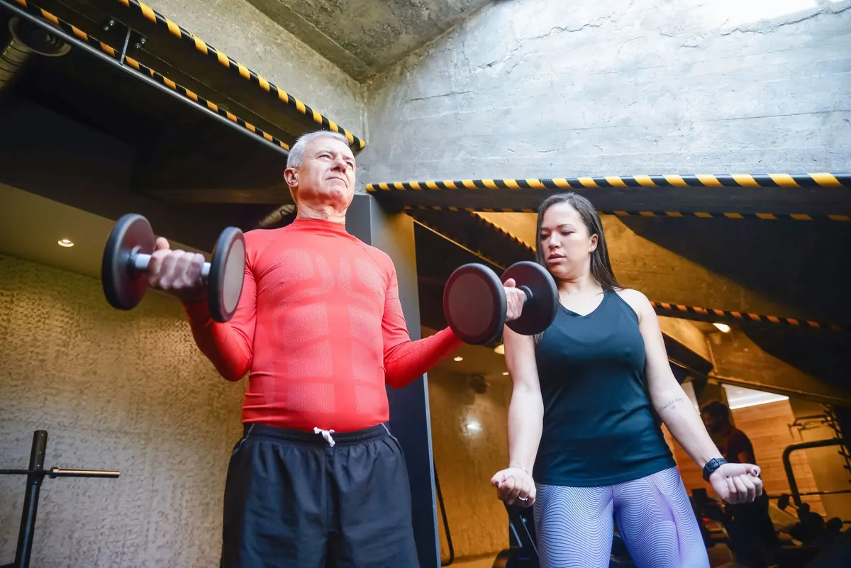 A personal trainer watches an older member's form as he curls a pair of dumbbells.